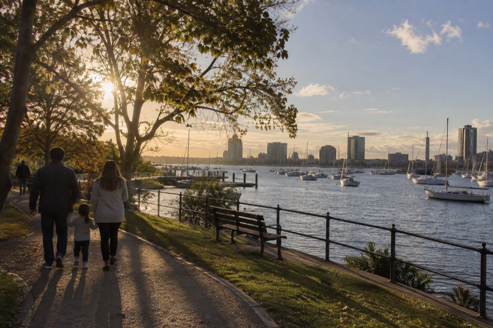 A family walking along a waterfront park path with sailboats moored in Boston Harbor behind them