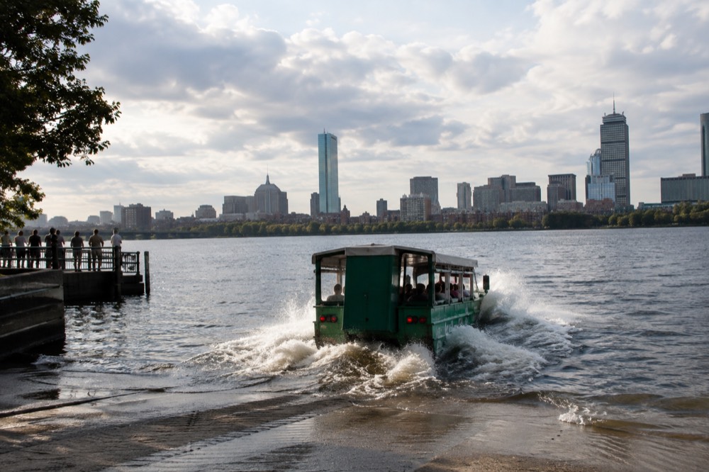 A duck boat vehicle splashing into the Charles River with the Boston skyline behind it