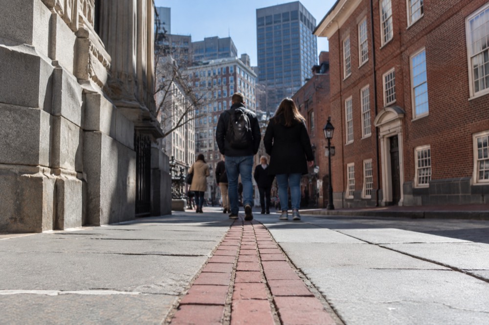 Visitors walking the red brick line of the Freedom Trail past historic Boston buildings on a sunny day