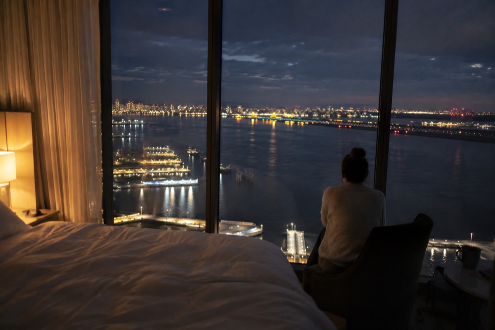 A hotel room with floor-to-ceiling windows facing the harbor and runway lights at night