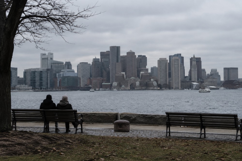 A waterfront park with benches facing the Boston skyline across the harbor channel