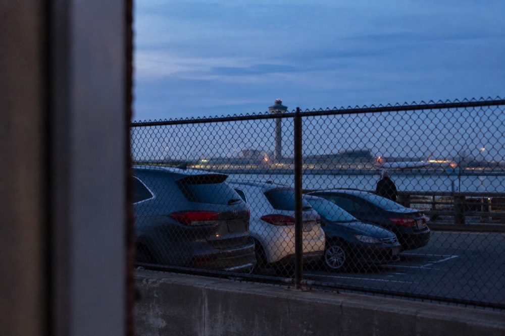 A row of parked cars in a fenced outdoor lot at dusk with an airport tower visible in the distance