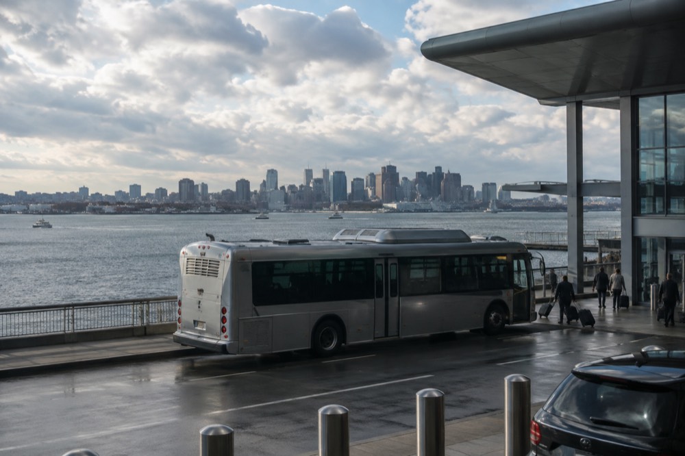 A silver bus at an airport curb with a city skyline across the water behind it