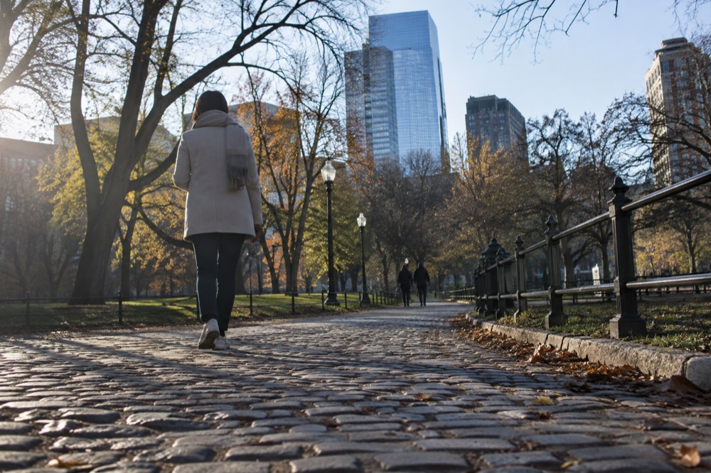 A person in a light jacket and scarf walking a cobblestone street in cool weather