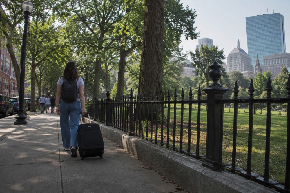 A traveler pulling a single carry-on suitcase down a sidewalk past brick row houses