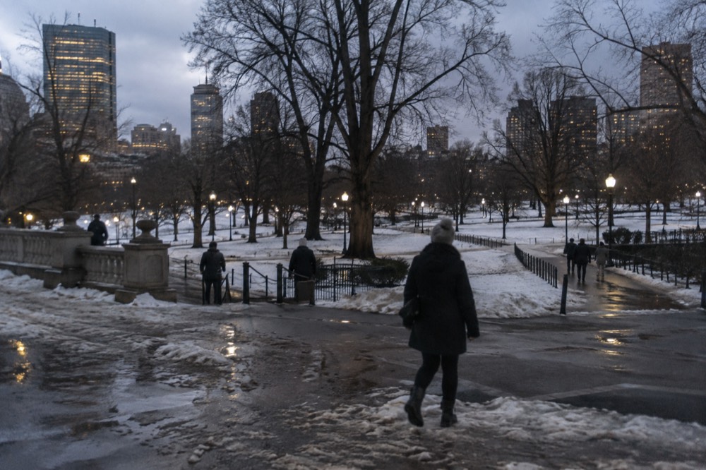 A walker in an insulated coat and boots crossing a slushy Boston sidewalk at dusk