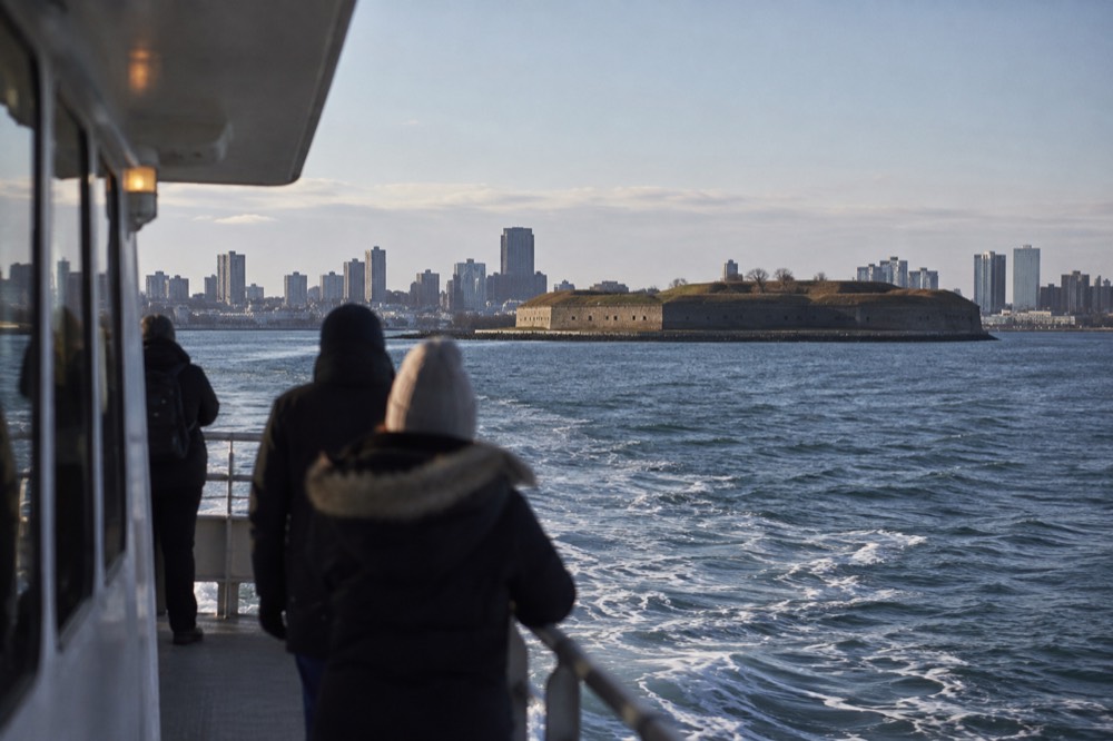 A ferry crossing open water toward a grassy harbor island with a stone fort