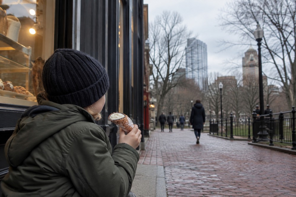 A child holding a cannoli outside a pastry shop on a narrow brick street
