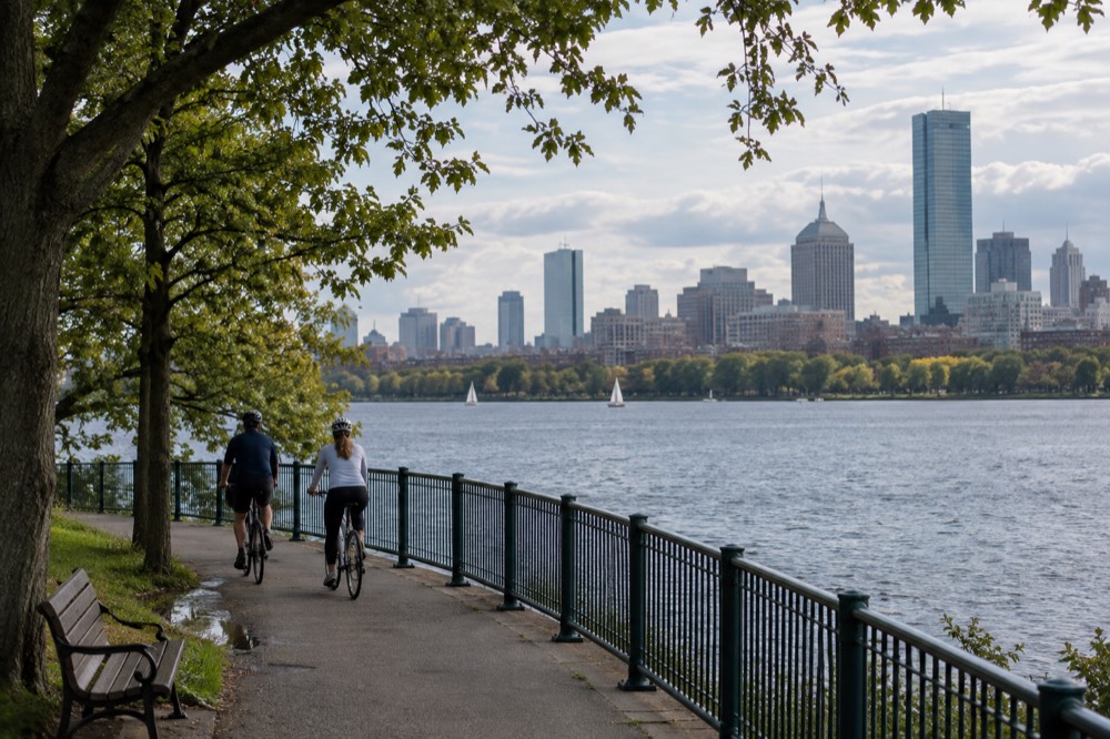 A couple riding bicycles along a riverside path with the Boston skyline behind them
